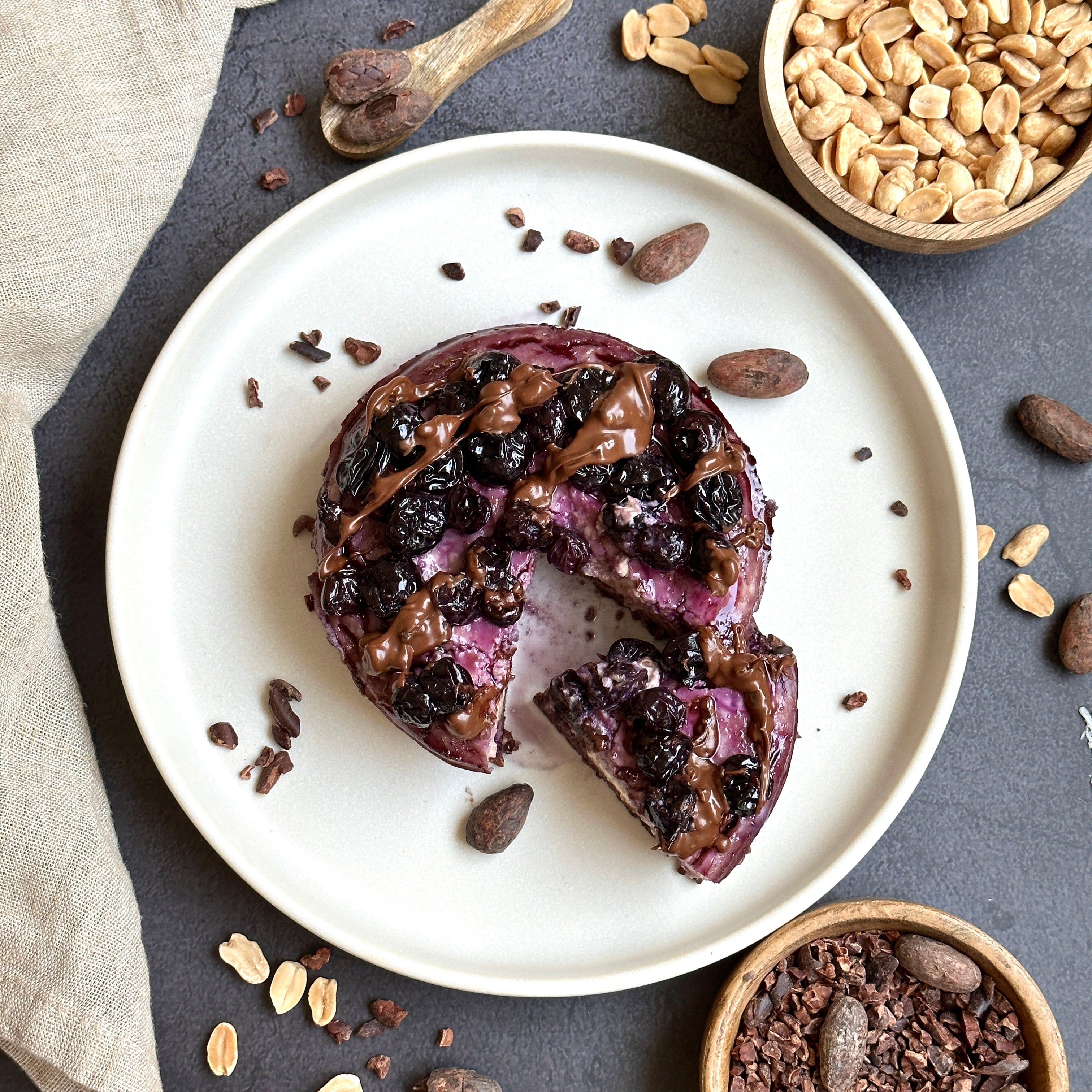 Proteinreicher Blaubeer-Cheesecake auf einem weißen Teller, getoppt mit Blaubeeren und dunklem Schoko-Protein-Spread.