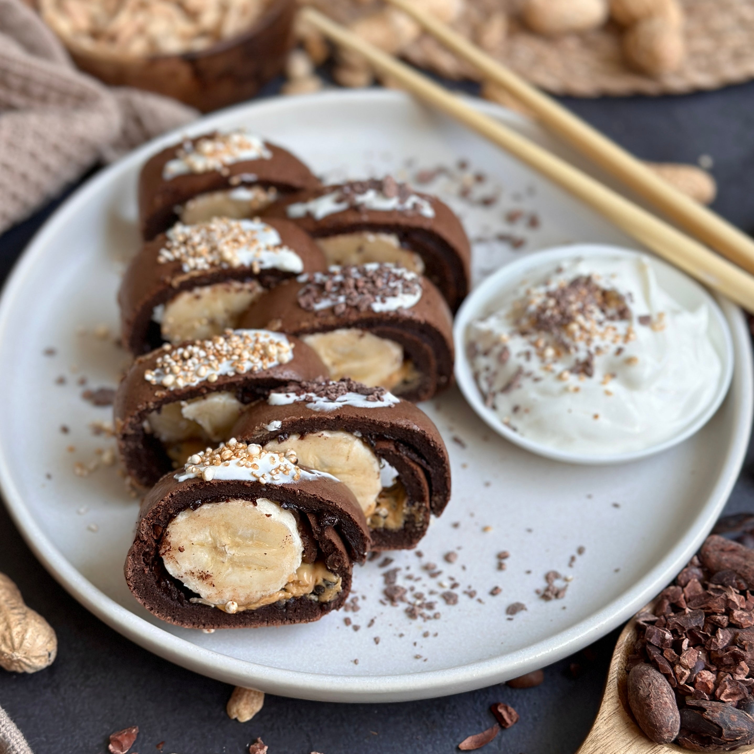 Erdnuss-Schoko Sushi mit Banane, Protein-Crêpe-Rolle in Sushi-Scheiben, serviert mit Joghurt-Topping und Kakao-Dekor.