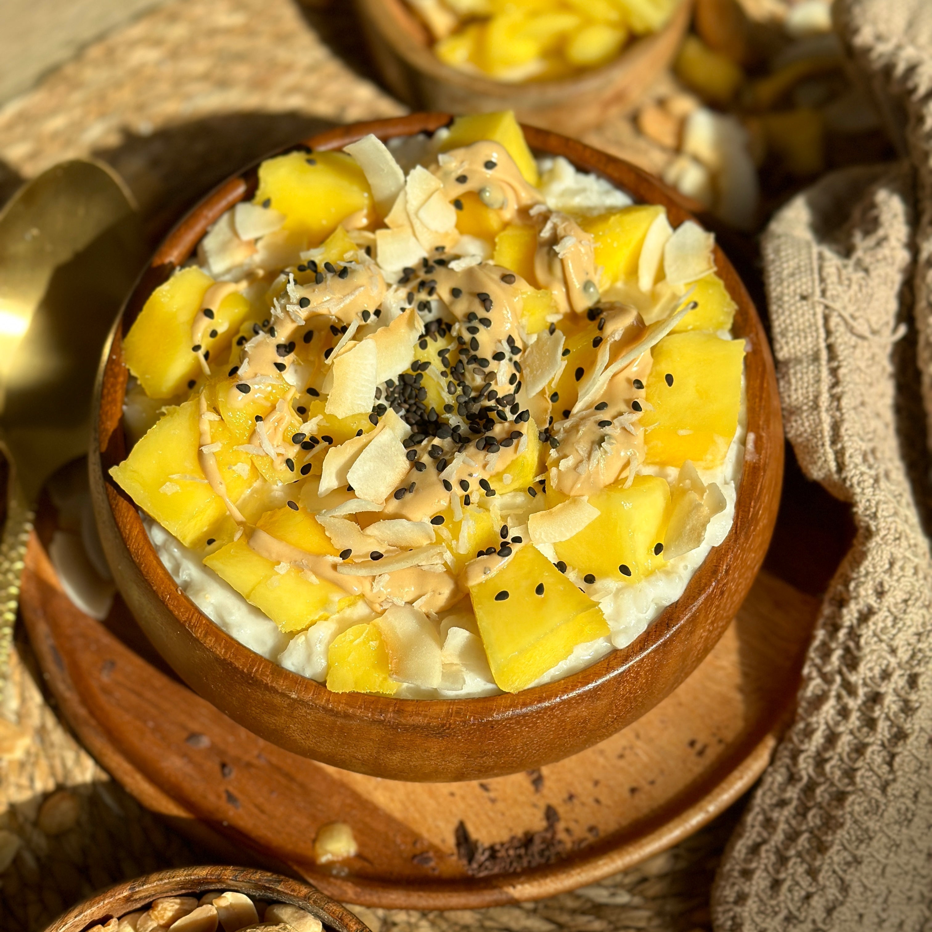 Schale mit cremigem Kokos-Mango Sticky Rice, garniert mit frischer Mango, schwarzem Sesam, Quinoa und einem Löffel Protein Erdnuss Spread.