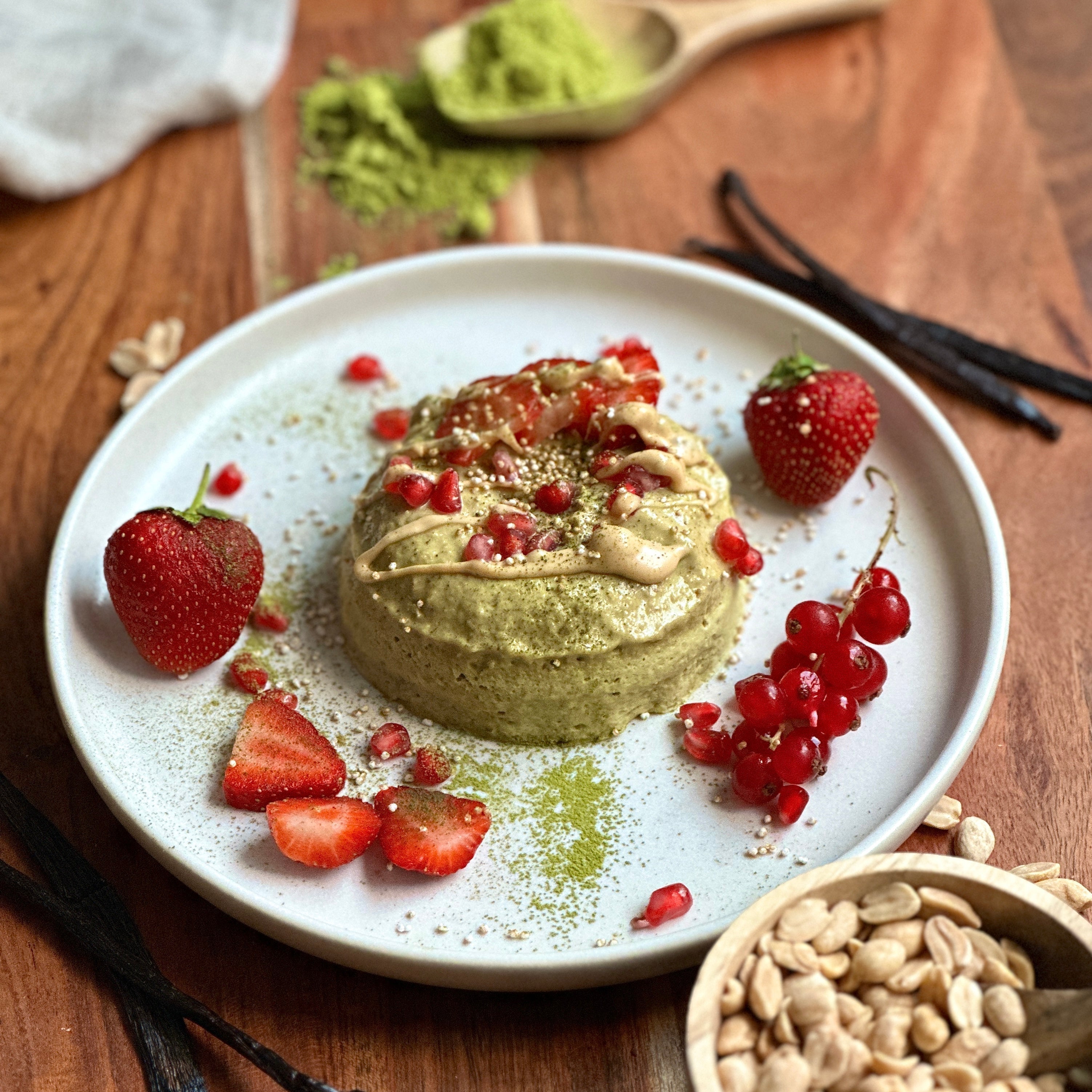 Saftiger Matcha-Mugcake mit cremigem Erdnusskern in einer weißen Schale, dekoriert mit Beeren und Granatapfelkernen.
